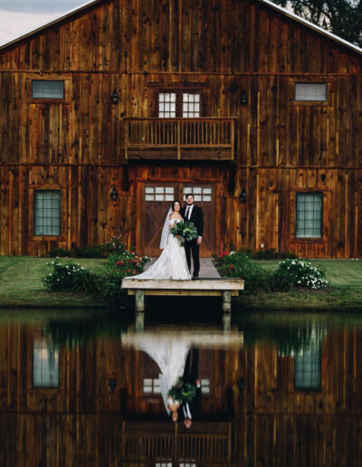 A bride and groom stand in front of the C Bar Ranch wedding barn with their images reflected in the pond.