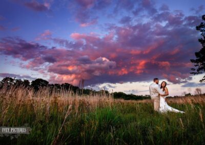 A newly married couple stands in a tree-lined field behind the wedding barn at C Bar Ranch in Alachua, Florida during a sunset of brilliant purple and pink clouds. Photo by Ivey Pictures of Jacksonville, Florida.