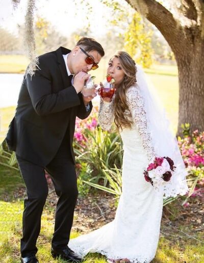 Bride and groom sip cocktails under a tree near pink blooming azaleas.