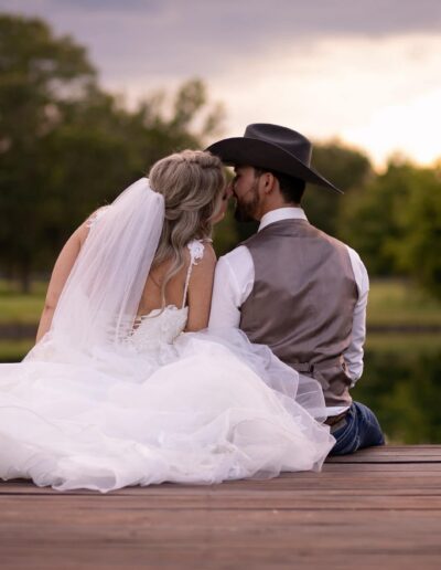 A bride and groom sit on dock at sunset.