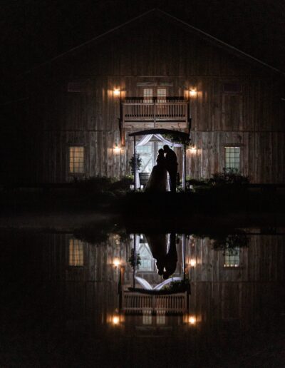 Bride and groom silhouetted in front of the C Bar Ranch barn at night with a reflected image in the pond.