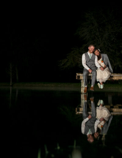 Bride and groom sitting on a dock with their image reflecting in the pond below.